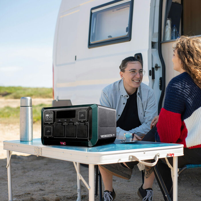 Two campers sit at a table chatting in front of a camper van at a beach campsite. On the camp table we see a green and black New Camping Power Stations From Welling & Crossley 
