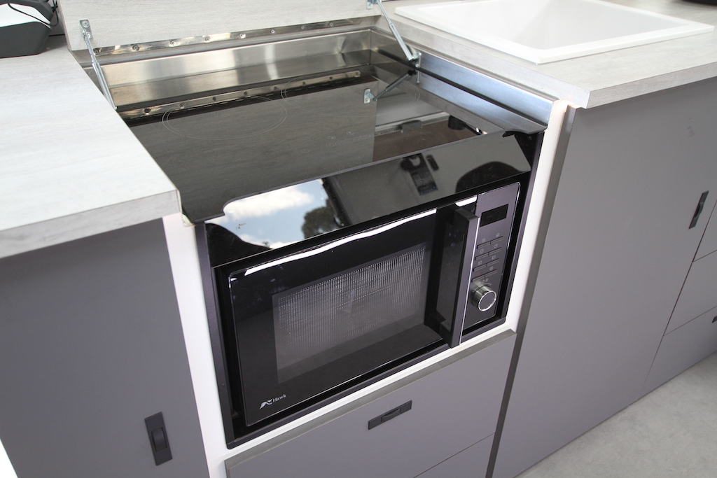 A close up of the multifunction 4-in-1 Microwave with Induction Cooktop built within cabinetry, just below benchtop height. A section of benchtop raised and supported with small gas struts reveals the top of the appliance, an induction cookop