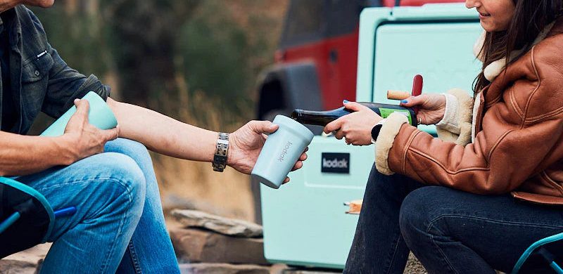 On camp chairs, a male camper holds out a Kodiak mug as a female pours a from a bottle of wine, in the background a mint coloured cooler that matches the mug