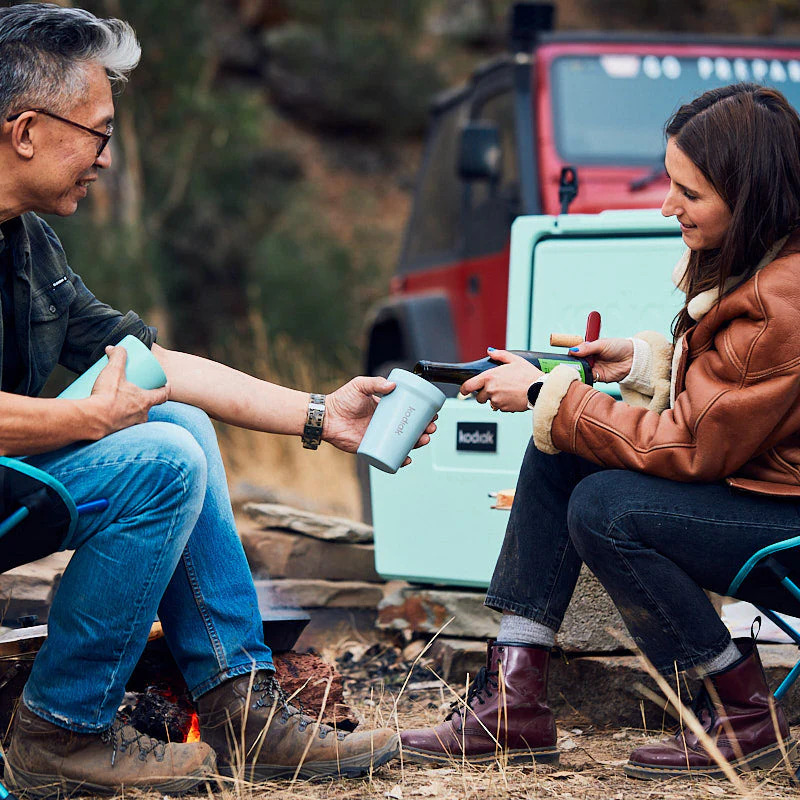 On camp chairs, a male camper holds out a Kodiak mug as a female pours a from a bottle of wine, in the background a mint coloured cooler that matches the mug